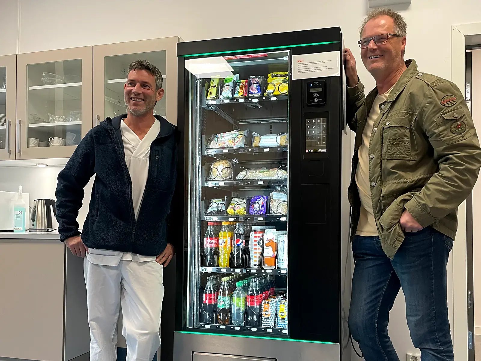 Vareautomat med mat og drikke på et kjøkken i Halden kommune, med to personer ved siden av